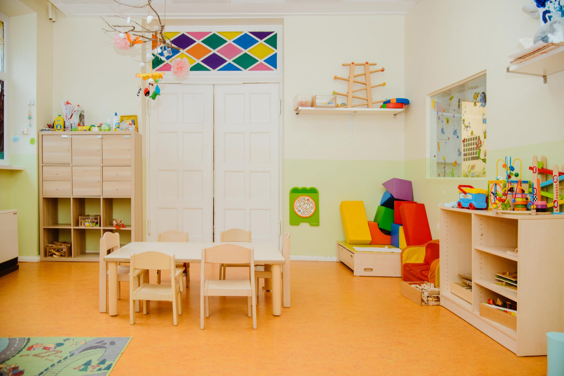 Heller Gruppenraum mit Kindertisch, Holzregalen, bunten Schaumstoffbausteinen und Fenster mit farbigem Rautenglas
